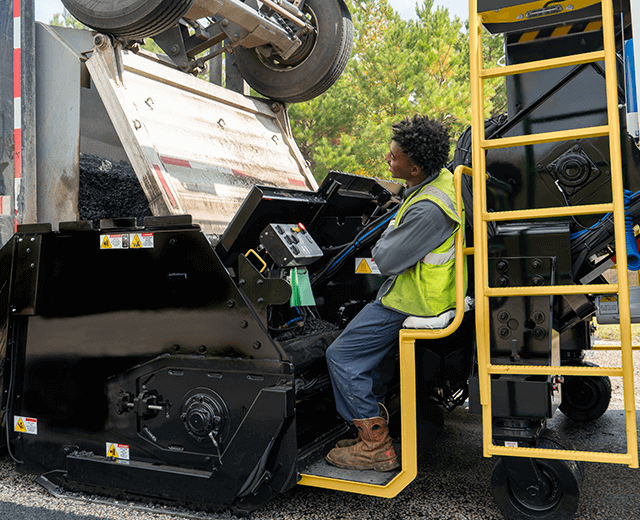 A Roadtec SB-3000 Shuttle Buggy Material Transfer Vehicle working as a dump truck unloads asphalt into the hopper with a ground crew memember sitting down at the ground control station